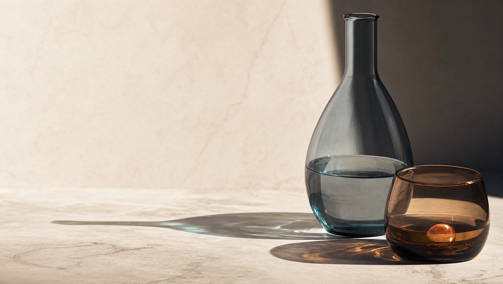 Two clean glass forms on a warm stone surface, one lit with a cool blue-grey highlight and one with a warm amber tone, half in shadow
