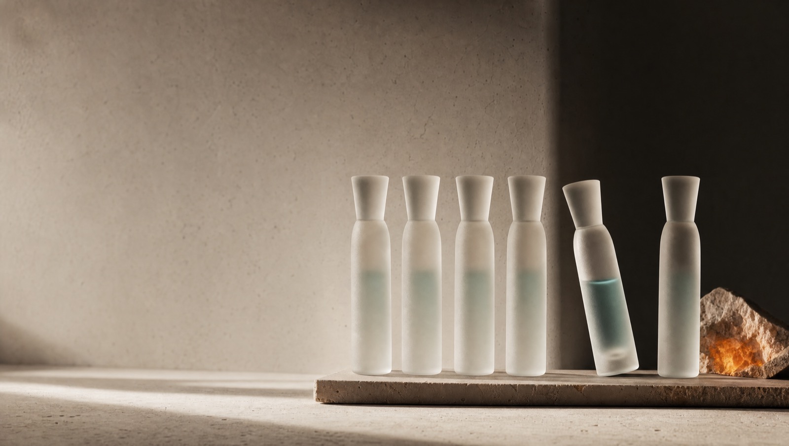 A row of evenly spaced frosted glass forms in a repeating rhythm on a warm stone surface, one subtly offset from the line, under soft side light